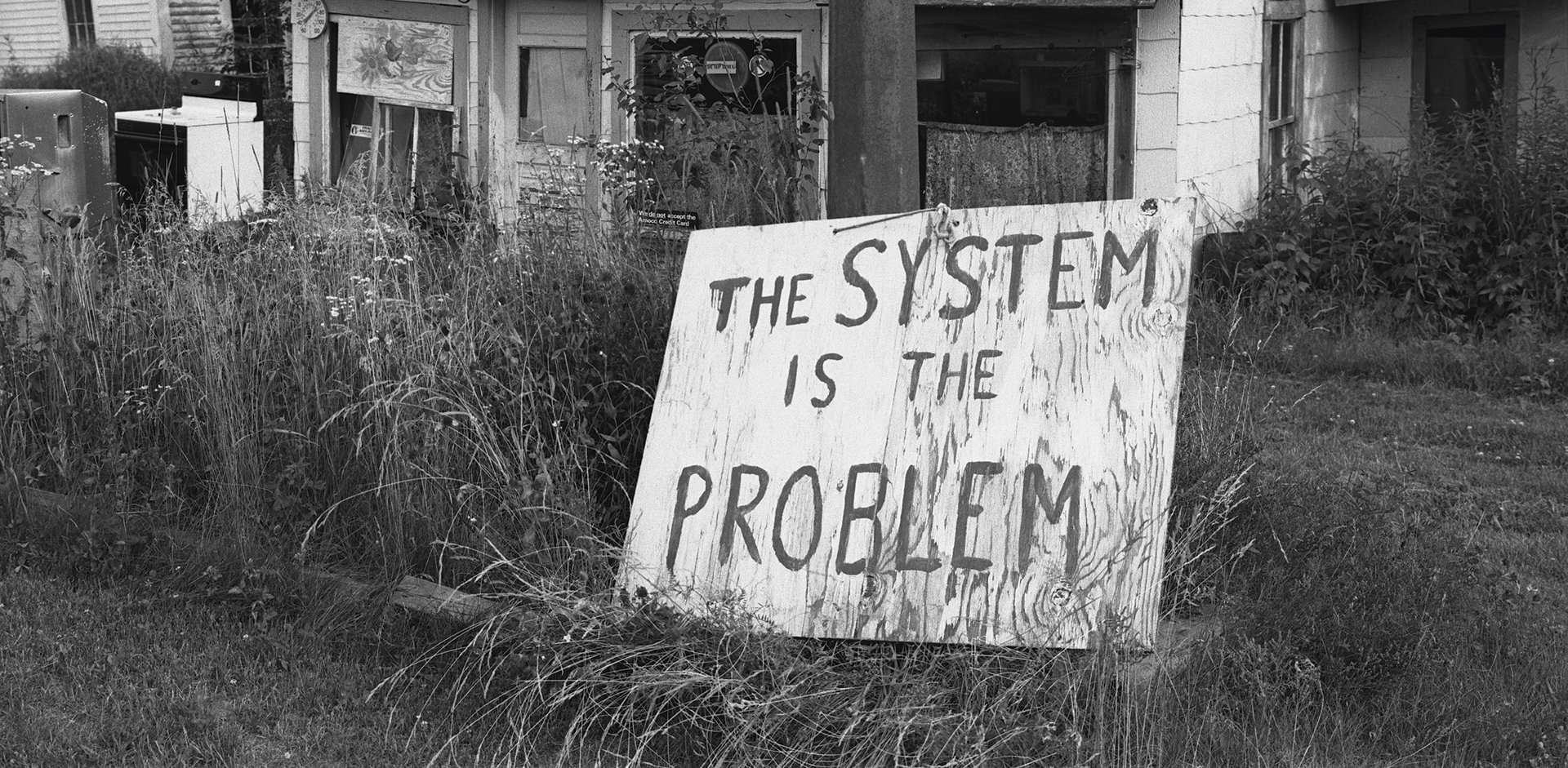 Black and white photo of ruined house with hand-painted sign "The System Is the Problem"