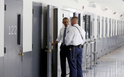 President Obama and a prison guard stand in front of an open door in a bright hallway, looking inside a prison cell.