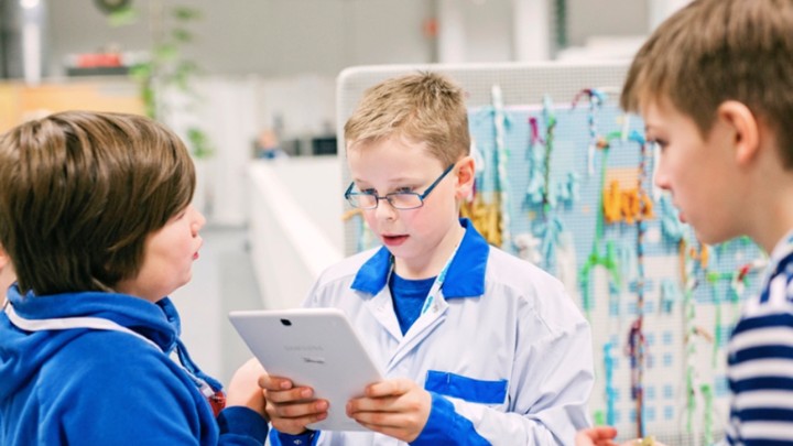 A sixth-grader stands in the center of the image looking at a hand-held electronic device and speaking to two other children in the frame.