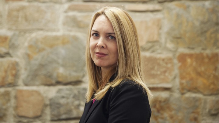 Larisa Dinsmoor, with shoulder-length blonde hair and light blue eyes, stands in front of a stone wall.