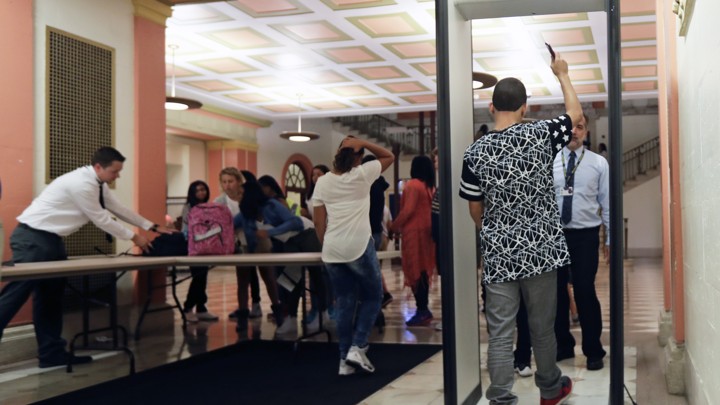 A student walks through a metal detector with his right arm in the air as other students collect their backpacks.