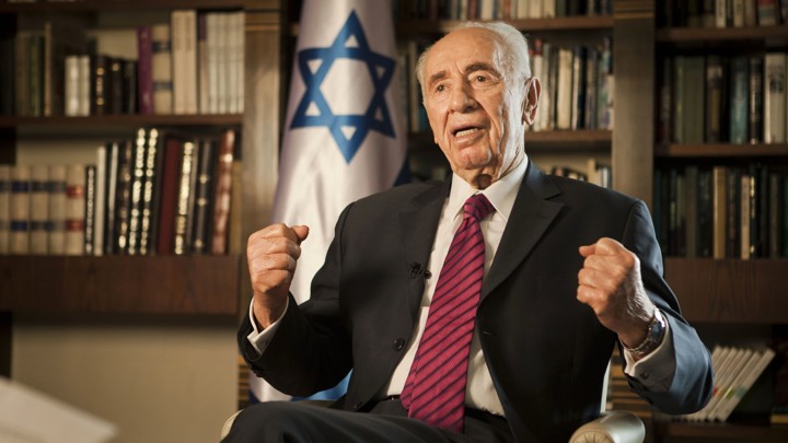 Shimon Peres gestures as he sits in front of bookshelves and an Israeli flag.