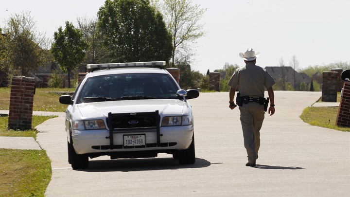 On a sunny day, a sheriff's deputy in a beige uniform, with his back to the photographer, walks alongside his gray squad card.