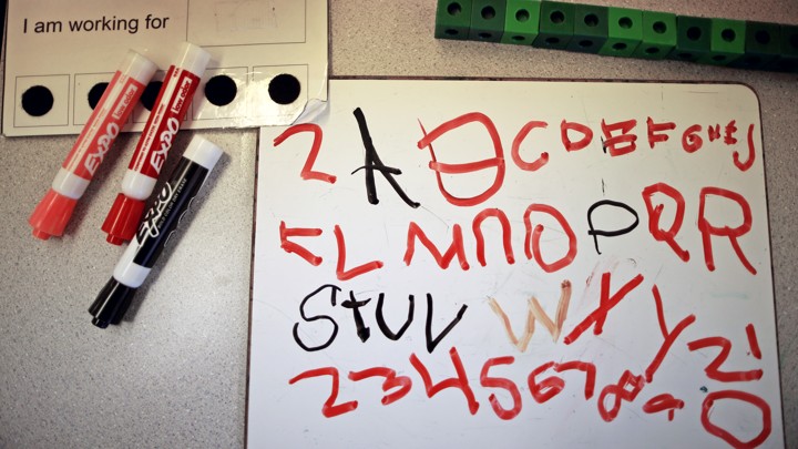 A whiteboard rests next to dry-erase markers and is covered in a student's attempts to scribble the alphabet.