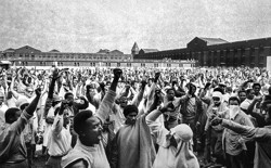 A black and white photograph depicts hundreds of inmates in the interior yard of Attica prison. Many have their hands raised in the "Black Power" gesture.
