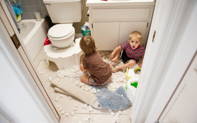 Two young children sit on the floor of a bathroom surrounded by packing peanuts. One child looks clearly alarmed and perhaps guilty of misbehavior.