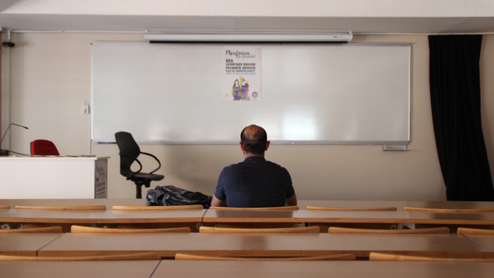 The back of a man sitting in an empty classroom, facing a blank dry-erase board