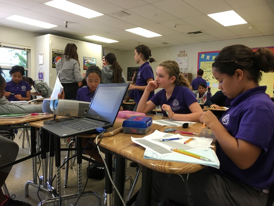Students dressed in purple polos sit in a classroom with papers, pencils, and a laptop strewn in front of them.