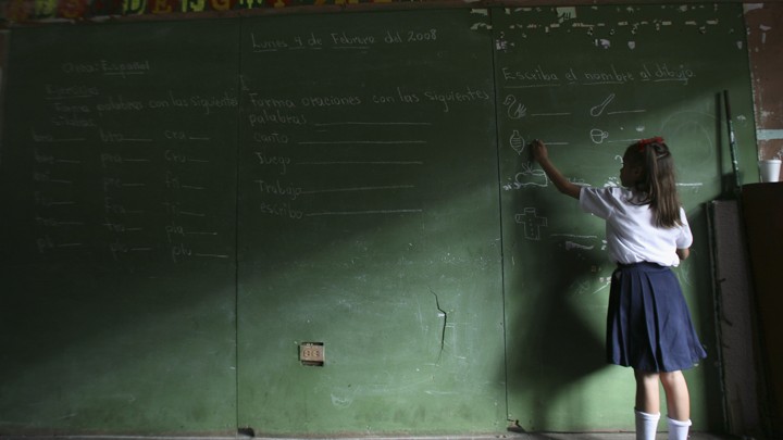 A child writes on a large chalkboard. Half the image is in shadows.