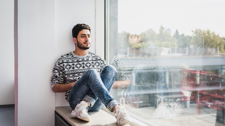 A young man sits on a ledge inside a building with a giant window.