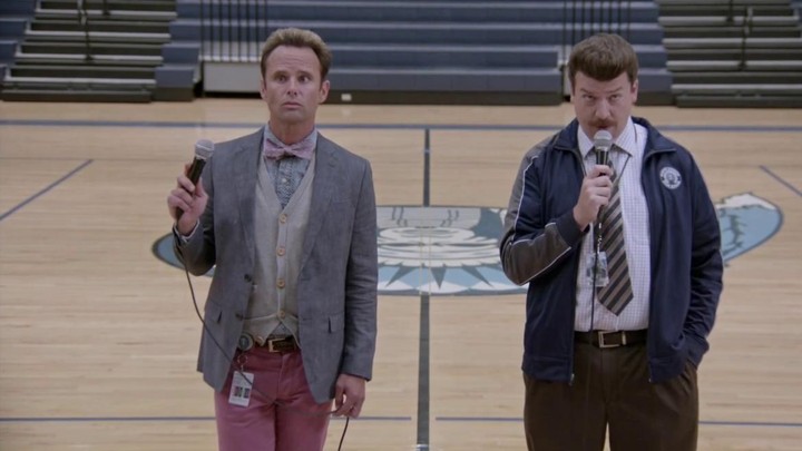 Walton Goggins, as Lee Russell, and Danny McBride, as Neal Gamby, stand in the center of a gymnasium holding microphones.