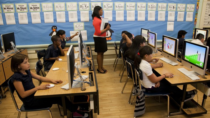A teacher stands at the back of a computer lab where students are seated at Mac desktop computers.