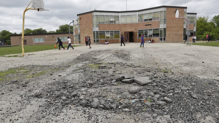 Kids play on a broken basketball court. The net on the hoop is missing. A brick school is in the distance.