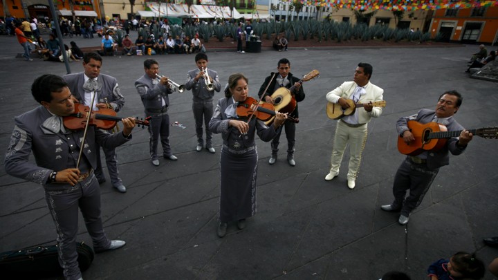 A mariachi band stands in a semi circle playing guitars, violins, and trumpets. A woman sands in the middle playing the violin.