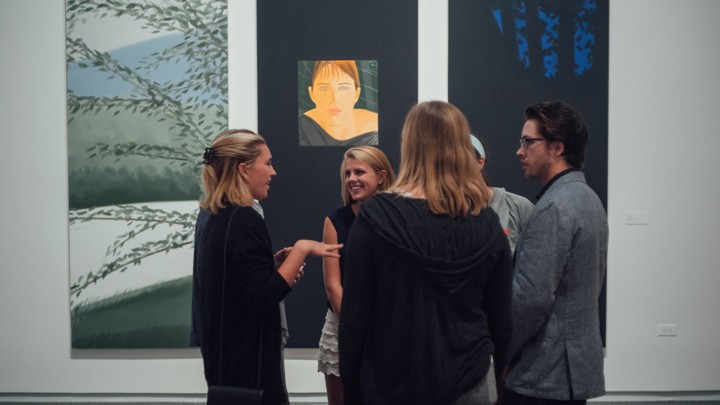 A group of five students stands, chatting, in front of three works of art: two paintings of tree branches and one portrait of a woman's face.