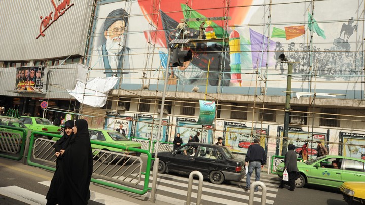 Young women fully covered in black chadors walk in front of a mural of Iran's leader Ayatollah Seyyed Ali Khamenei being refurbished on the corner of Revolution Square on March 1, 2012 in Tehran, Iran.