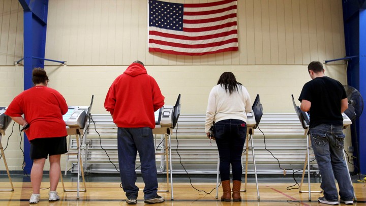 The backs of voters as they cast their ballots. They stand underneath an American flag, presumably in a gymnasium.