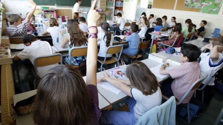 A classroom full of students listens to a teacher in front of a chalkboard. Two students' hands are raised.