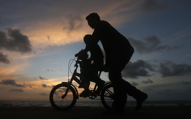 A man guides a child riding a two-wheel bike. It's sunset, and the figures appear as silhouettes.