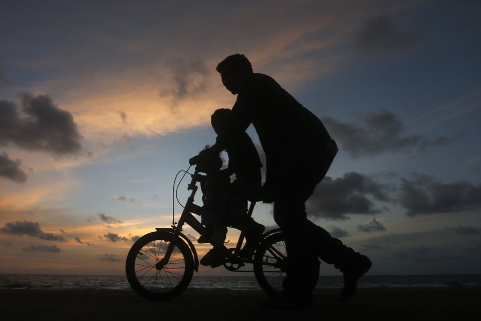 A man guides a child riding a two-wheel bike. It's sunset, and the figures appear as silhouettes.