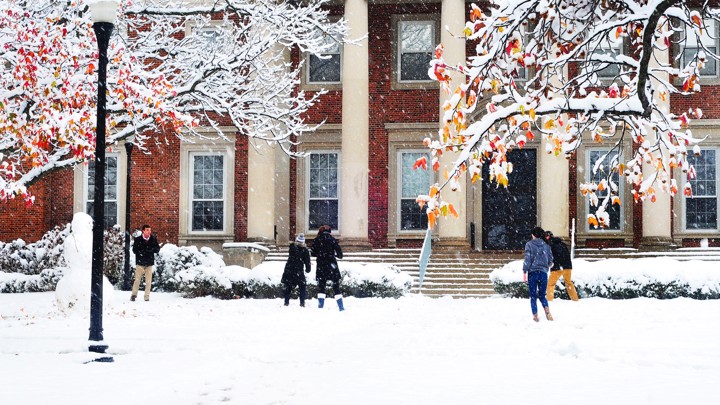Students have a snowball fight in front of a large brick building with columns.