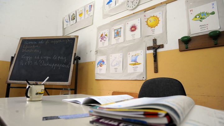 A teacher's desk is covered with papers in a classroom with a chalkboard and crucifix.