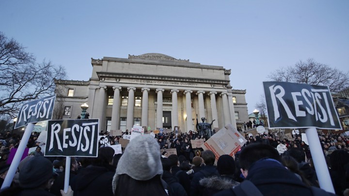 Students hold signs reading "resist" outside a stately university building with grand columns.