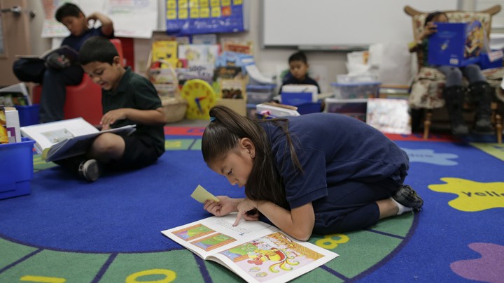 A girl crouches over a book on a colorful rug. Other children around her also have books on their laps.