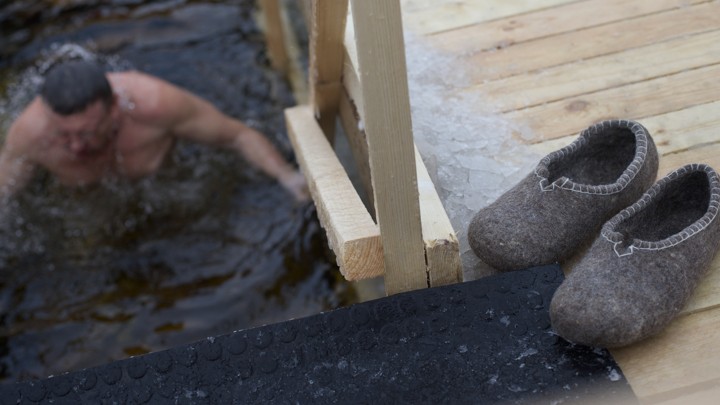 A Russian man eases himself into icy water while his slippers wait on a deck.