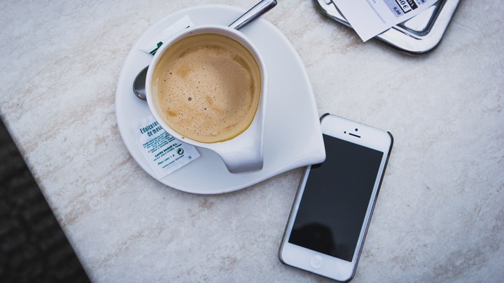 A smartphone sits unattended by a cup of coffee on a table.