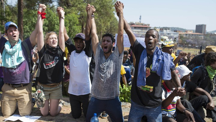 Five young adults sit on their knees, holding hands with their arms above their heads. Their mouths are open as they appear to be yelling.