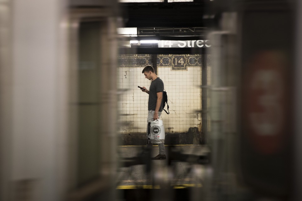 Seen between the cars of a passing train, a man looks at his phone on a subway platform.