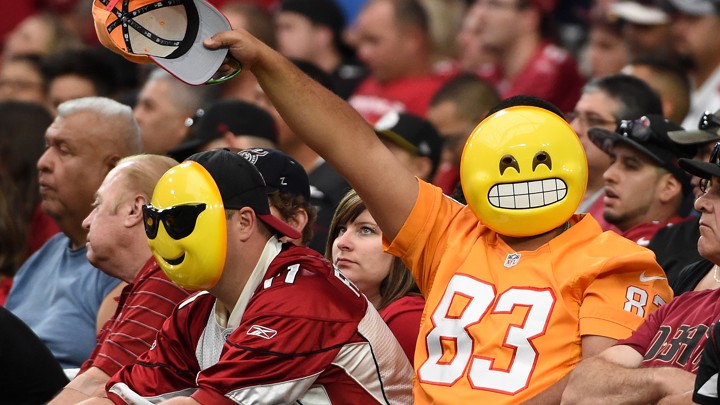 Fans wearing emoji masks wave from the stands at an NFL game.
