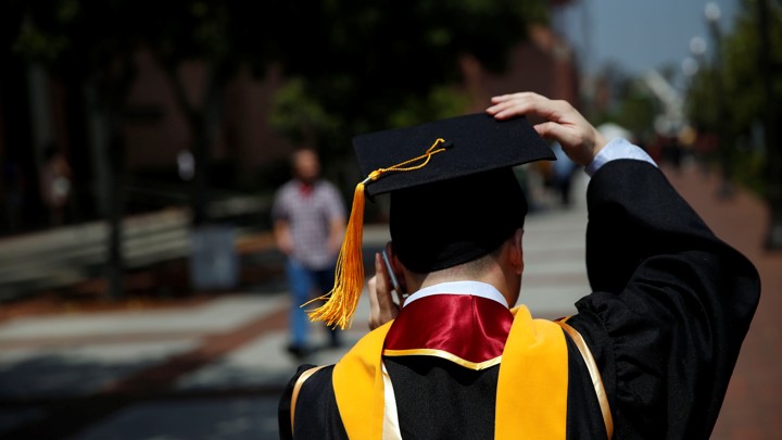 The back of a graduation in a cap and gown. He is talking on his cell phone.