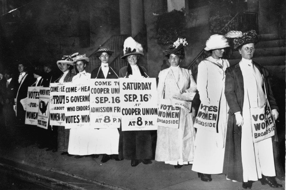A black and white photo of women in dresses and feathered hats. They hold signs advertising an upcoming rally for women's right to vote.