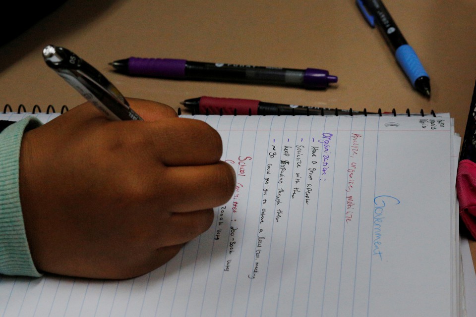 A student holds a pencil above a sheet of notebook paper. The notes appear to be about the government