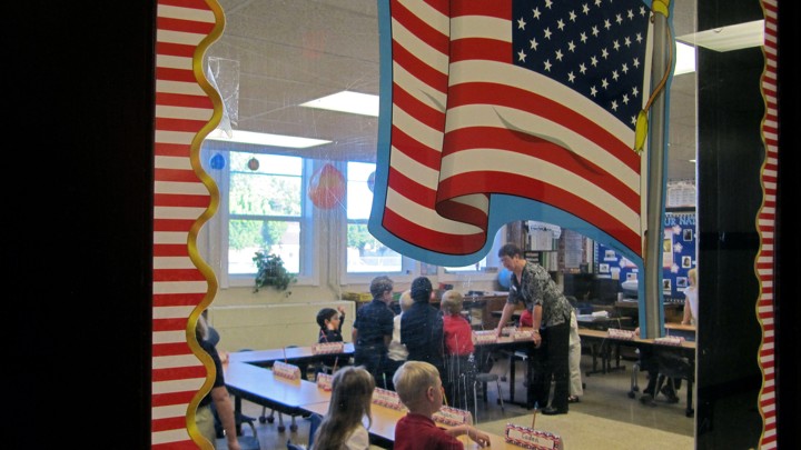 The photograph peers into an elementary school classroom through a door with an American flag sticker on it.
