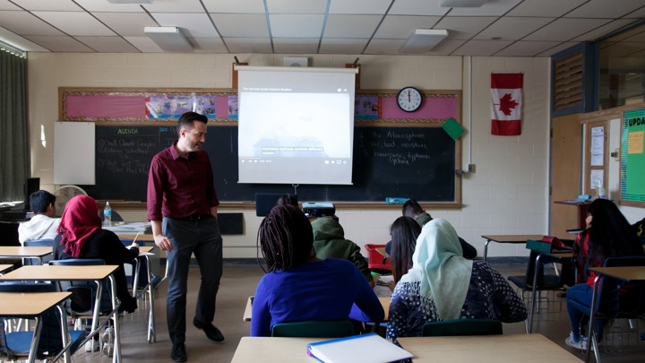 A teacher stands at the front of a classroom in Canada; students sit at desks, a few of whom are wearing hijabs, one of whom has dreadlocks.