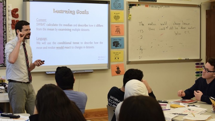 A teacher stands near a whiteboard that describes "Learning Goals" and another board with math problems; students sit at desks.