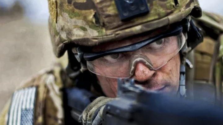 A person in camouflage clothing and helmet, wearing safety goggles, peers down the barrel of a gun.