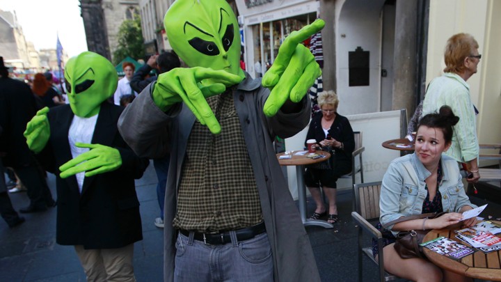 Two people dressed in green alien heads and hands gesture on a busy street.