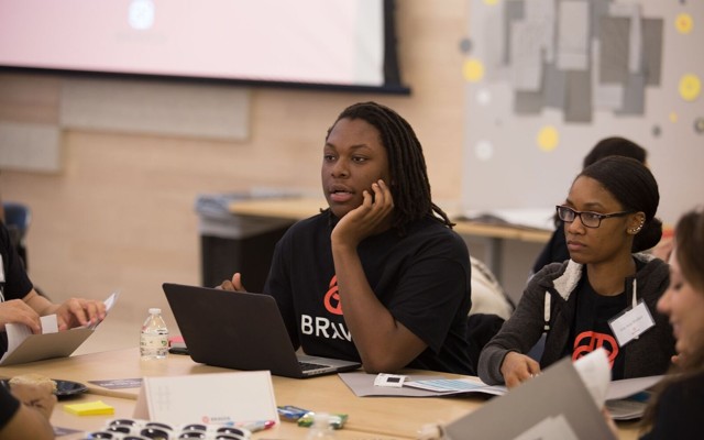 Two students sit at a desk, one of them working at a computer. 