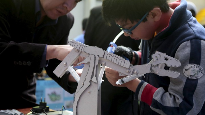 A boy builds a dinosaur robot; his teacher assists him.