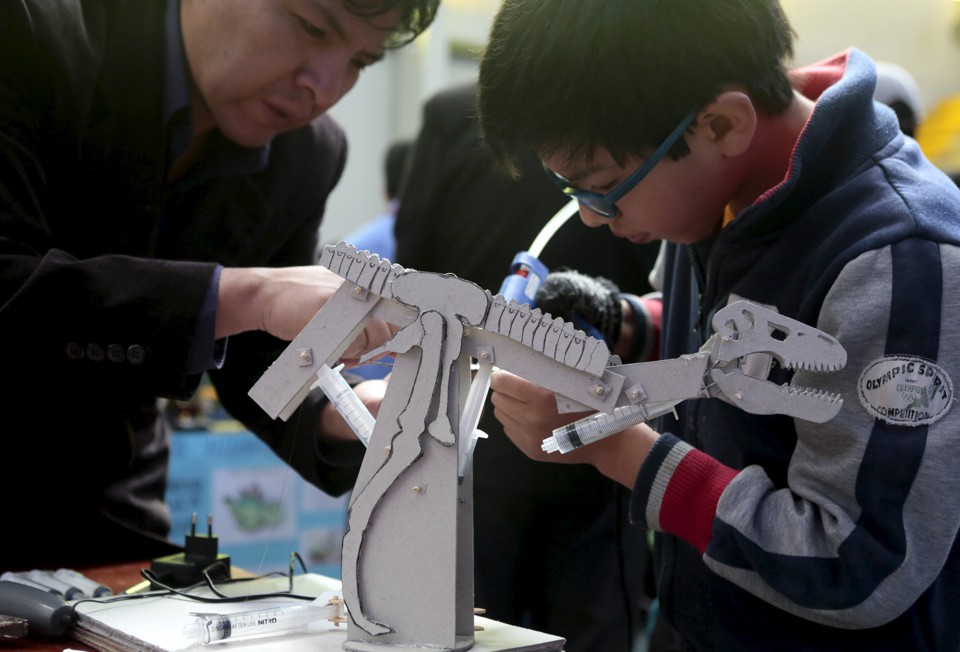 A boy builds a dinosaur robot; his teacher assists him. 