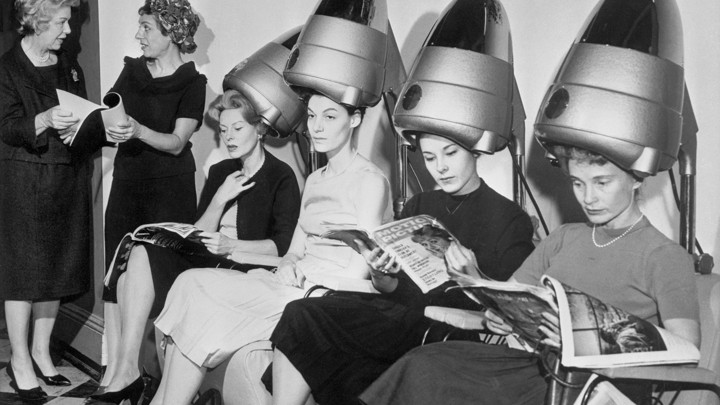 A group of women sit underneath hair dryers and read magazines at a hair salon.