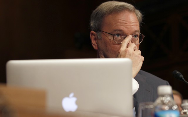 A man sitting behind an Apple laptop covers the lower half of his face with his hand.