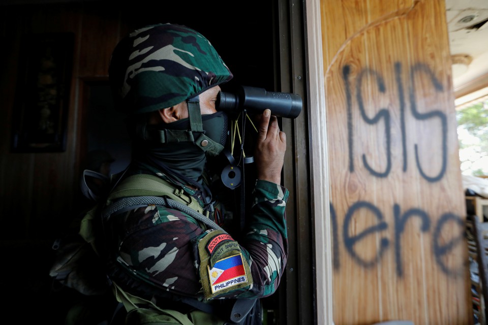 A Filipino soldier uses binoculars next to a wood panel where the words ' ISIS here' are spray-painted.