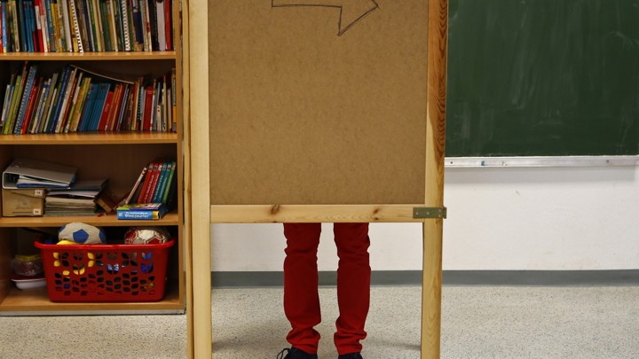 A pair of legs are visible behind a wooden corkboard in a classroom.