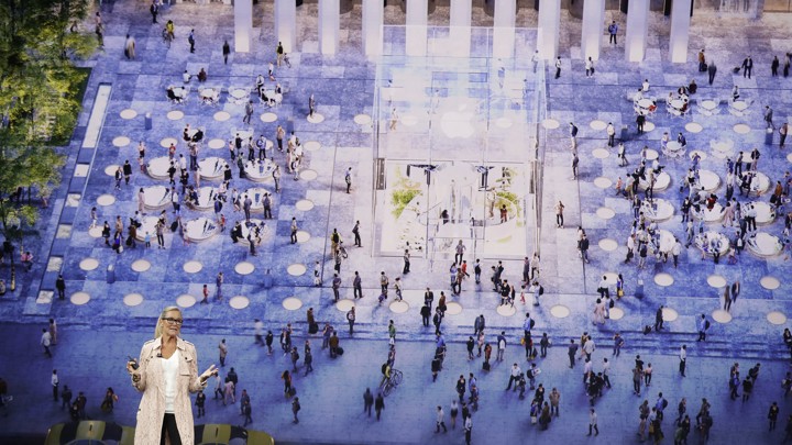 An Apple speaker stands in front of a screen depicting people gathered in a town square