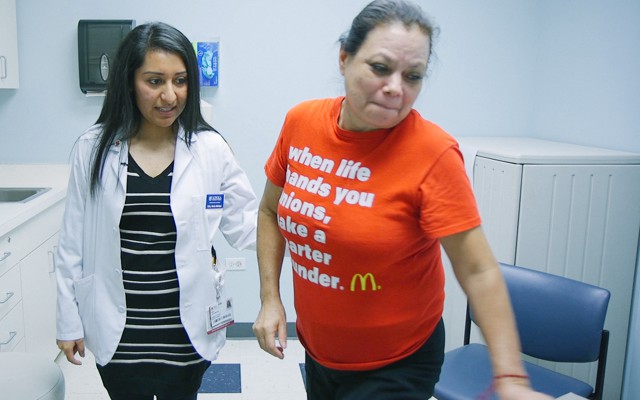 A woman in a lab coat supports a woman in a T shirt in a doctor's office.